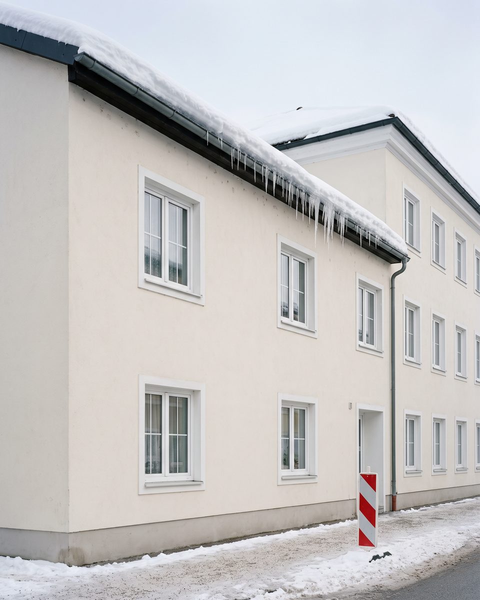 Schneewächte und Eiszapfen an der Dachrinne über einem geräumten Gehsteig, eine rot-weiße Warnbake am Gehsteigrand